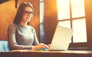 Frau mit langen braunen Haaren und Brille sitzt vor ihrem Laptop und lächelt zufrieden
