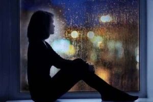 Aufnahme einer Frau, am Fenster sitzend, sie schaut traurig und nachdenklich auf die verregneten Straßen