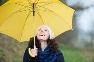 Aufnahme einer fröhlichen, gesunden Frau die mit einem leuchtend-gelben Regenschirm dem Herbstregen trotzt