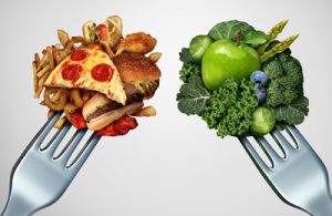 Zwei Gabeln - auf der linken Gabel sind ungesunde, fettreiche Lebensmittel aufgepiekst und auf der rechten sieht man gesundes Gemüse und Obst